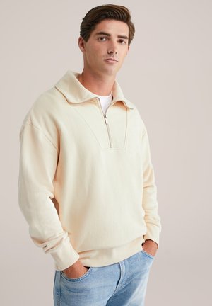 Crème-kleurige zip-up sweatshirt gemaakt van zachte stof, met een brede kraag en een relaxte pasvorm. Draagt over lichtblauwe jeans.