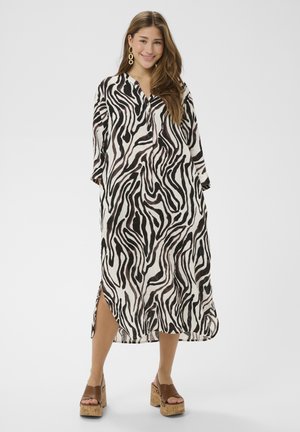 Tânără purtând o rochie midi cu imprimeu de zebră negru și alb, cu nasturi, cercei aurii și sandale maro cu platformă.