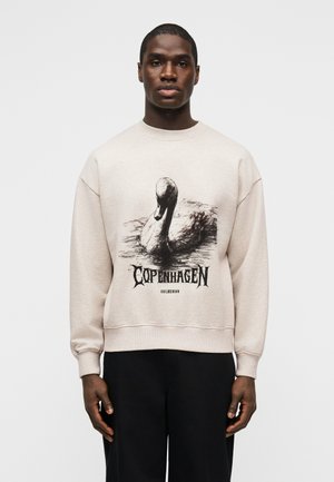 Mann trägt beige Sweatshirt mit schwarzem und weißem Schwan-Motiv und "Copenhagen"-Schriftzug, kombiniert mit schwarzer Hose, steht vor weißem Hintergrund.