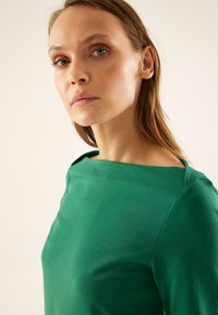 Groene top met een gladde, aansluitende pasvorm, voorzien van een bootnek en subtiele schouderaccenten. De stof komt zacht en licht aan.