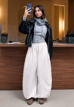 Fiatal nő fekete bőrdzsekiben, szürke kapucnis pulóverben, széles fehér nadrágban és sportcipőben, aki szelfit készít egy modern, fa pultokkal berendezett beltéri helyszínen.