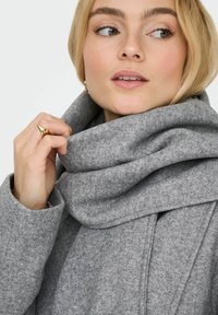 Cappotto di lana grigio con colletto drappeggiato e texture morbida, caratterizzato da un design a piega. Il modello tiene una mano al colletto. Accento con anello dorato.