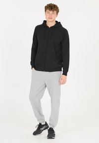 Mustat vetoketjuhupparit, joissa on säädettävät nyörit ja etutaskut, yhdistettyinä vaaleanharmaisiin collegehousuihin ja mustiin urheilukenkiin. Sujuva rakenne.