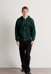 Mørkegrøn zip-up hoodie med en tætsiddende design, ribbede håndled og en kengurulomme, parret med sorte bukser og sko. Simpel, tekstureret stof.