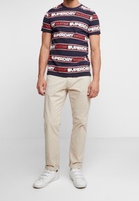 Marineblå stribet t-shirt med rød og hvid "SUPERDRY TRACKSTARS" tekst, parret med beige bukser og hvide sneakers. Casual fit, lavet af bomuldsmateriale.