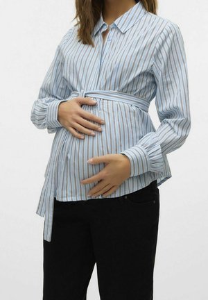 Zwangere vrouw die een blauw-wit gestreept overhemd met knopen draagt en met beide handen haar buik vasthoudt, gecombineerd met een zwarte broek.