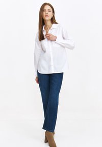 Biała koszula button-up z klasycznym kołnierzykiem, długimi rękawami, w zestawieniu z ciemnoniebieskimi spodniami typu straight-leg i beżowymi botkami na obcasie.