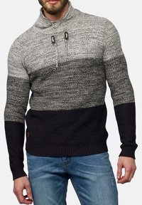 Man som bär en svart och grå stickad tröja med hög krage och knappar, tillsammans med blå denimjeans.