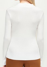 Haut blanc à manches longues côtelé avec une coupe près du corps, doté d'un col arrondi et d'une texture subtile, associé à un pantalon marron.