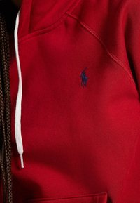 Felpa rossa con cappuccio realizzata in tessuto misto cotone, con un laccetto bianco e un piccolo logo blu navy sul petto. Texture liscia, tasca a marsupio.