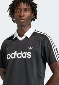 Μαύρη φανέλα Adidas με λευκές ρίγες και γιακά, με μεγάλο λευκό λογότυπο. Το ύφασμα φαίνεται λείο με διακριτική γυαλάδα.