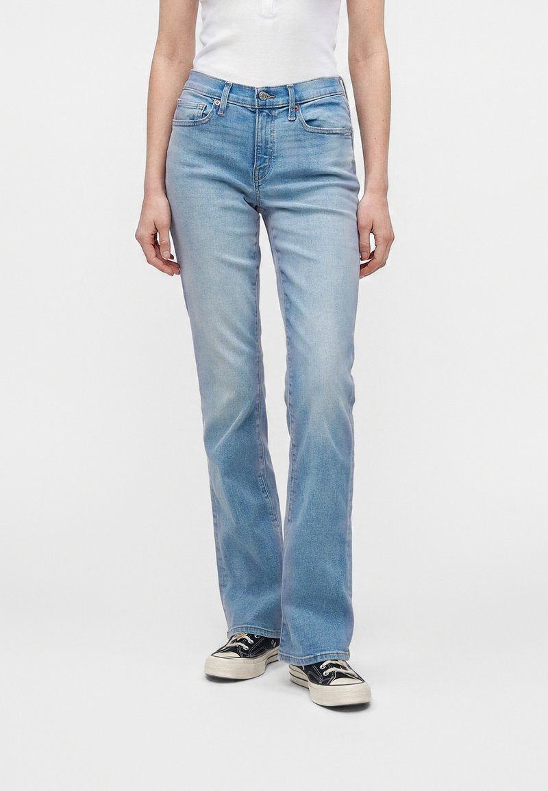 Γυναίκα με ανοιχτό μπλε τζιν κοπής bootcut, λευκό αμάνικο τοπ και μαύρα χαμηλά αθλητικά παπούτσια με λευκές σόλες και κορδόνια.