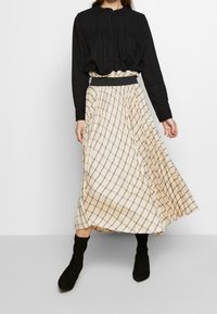 Svart blus med lång ärm och veck, parat med en knälång, beige kjol med rutmönster; modellen bär svarta, mockastövlar.