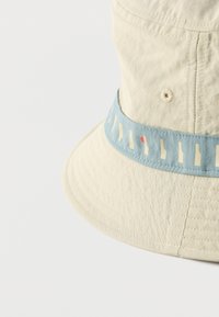 Μπεζ καπέλο τύπου bucket με μια γαλάζια ταινία που διαθέτει αφηρημένα κρεμ σχήματα και μια μικρή κόκκινη κουκκίδα γύρω από την κορυφή.