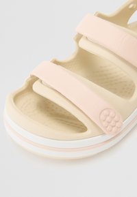 Sandalia de un beige claro con una superficie texturizada, que cuenta con dos tiras anchas de color rosa y un acento redondo. La suela tiene una combinación de colores en blanco y rosa.