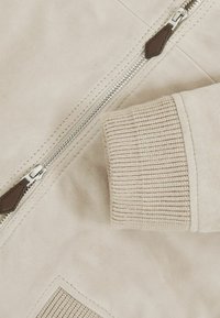 Heled beez suede jakk, millel on hõbedane tõmblukk, soonikkoes varrukad ja tekstuuriga detail varrukal.