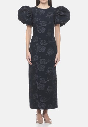 Donna che indossa un lungo vestito nero con maniche corte a sbuffo e motivo floreale scuro, abbinato a tacchi a punta color nude.