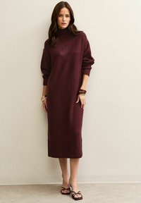 Donna che indossa un lungo vestito in maglia bordeaux con maniche lunghe e sandali neri con punta aperta, in piedi contro un muro beige.
