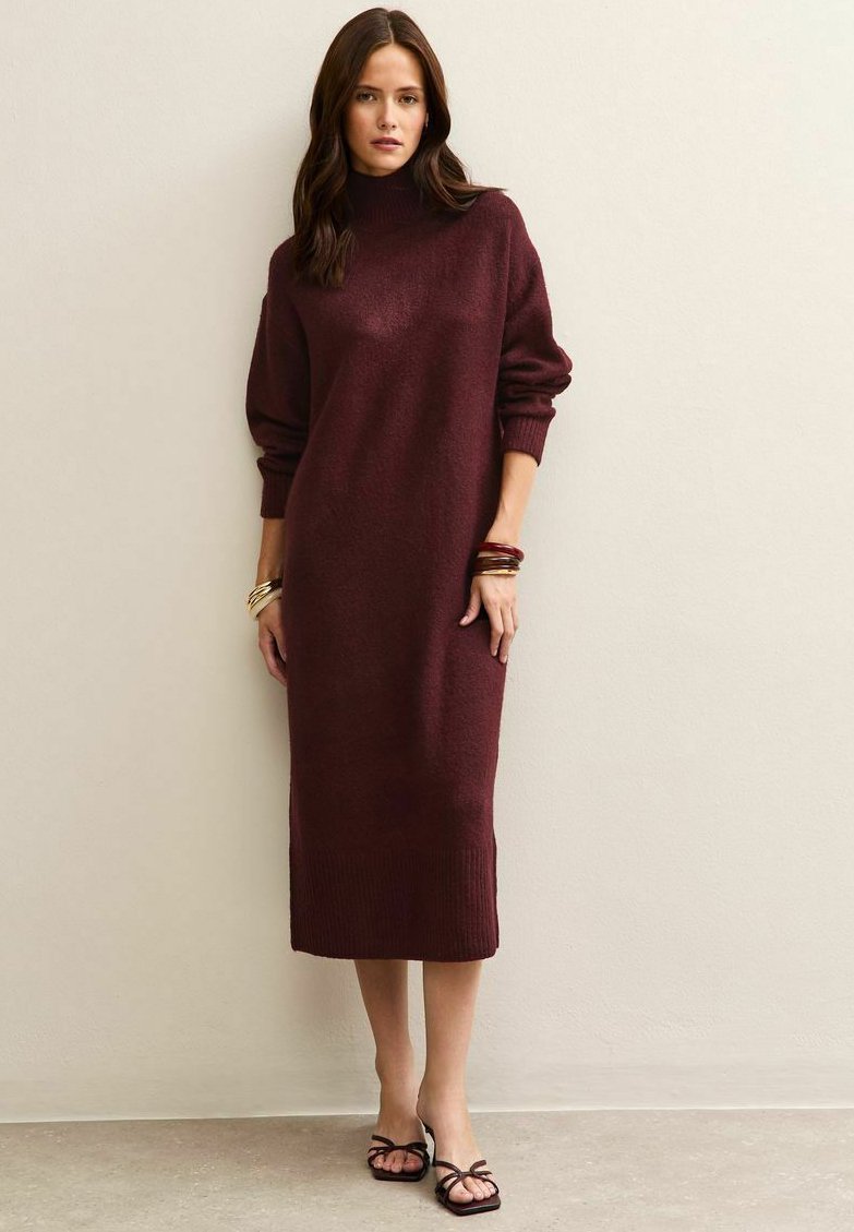 Donna che indossa un lungo vestito in maglia bordeaux con maniche lunghe e sandali neri con punta aperta, in piedi contro un muro beige.