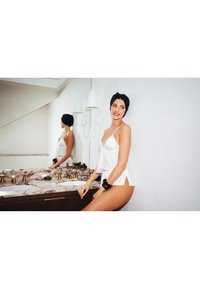 Set di lingerie bianco con pizzo, composto da una maglietta ampia e fluida e pantaloni coordinati. Sullo sfondo si trova un vanity in marmo con hardware dorato.