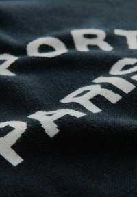 Ciemnoniebieska tkanina z wyraźnym białym napisem, o miękkim, teksturowanym wyglądzie. Litery mają wyraźny, lekko nieregularny kształt.