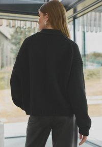 Juodas megztukas su laisvu siluetu, su raukšlėtu apvadu ir rankogaliais, priešais stiklinę foną. Lygus audinys ir subtilūs detalės.
