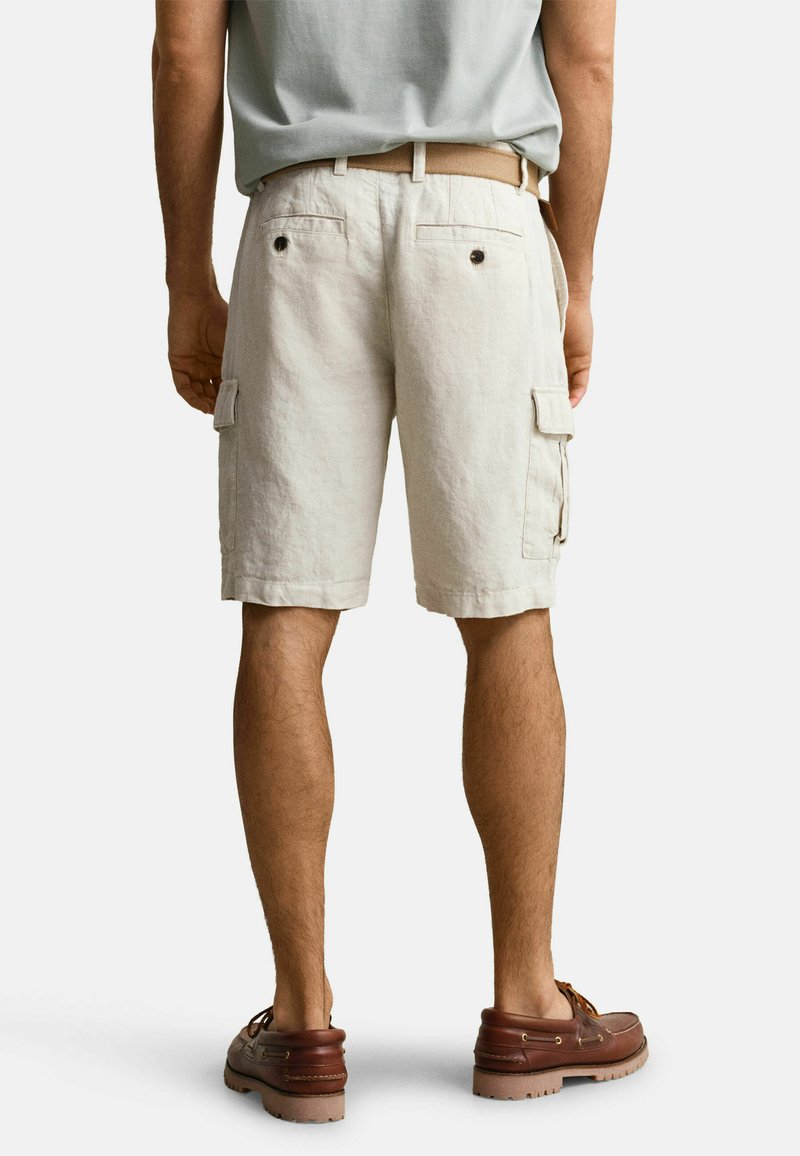 Hombre que lleva pantalones cortos cargo beige con bolsillos laterales y traseros, un cinturón claro, camisa gris claro y zapatos de cuero marrón, de pie sobre un suelo blanco.