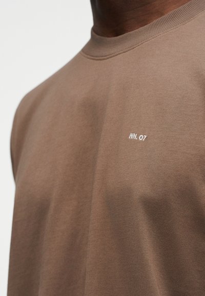 Κοντινή εικόνα ενός καφέ μπλουζάκι με στρογγυλή λαιμόκοψη και μικρό λευκό κείμενο "NN.07" στο αριστερό στήθος, που φοράει ένα άτομο με σκούρο χρώμα δέρματος.