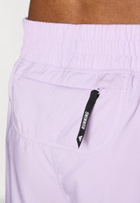 Shorts de course violet clair avec une taille élastique et une petite poche zippée dotée d'une languette noire étiquetée "COURSE". Texture lisse.