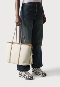 Beige canvas tote bag with tan leather accents, rectangular shape, and a decorative charm. Worn with high-waisted blue denim and athletic shoes.