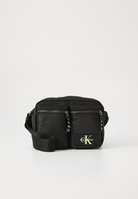 Black nylon crossbody bag with two front zip pockets, adjustable strap, and a yellow logo patch. Features a sleek and minimalist design.