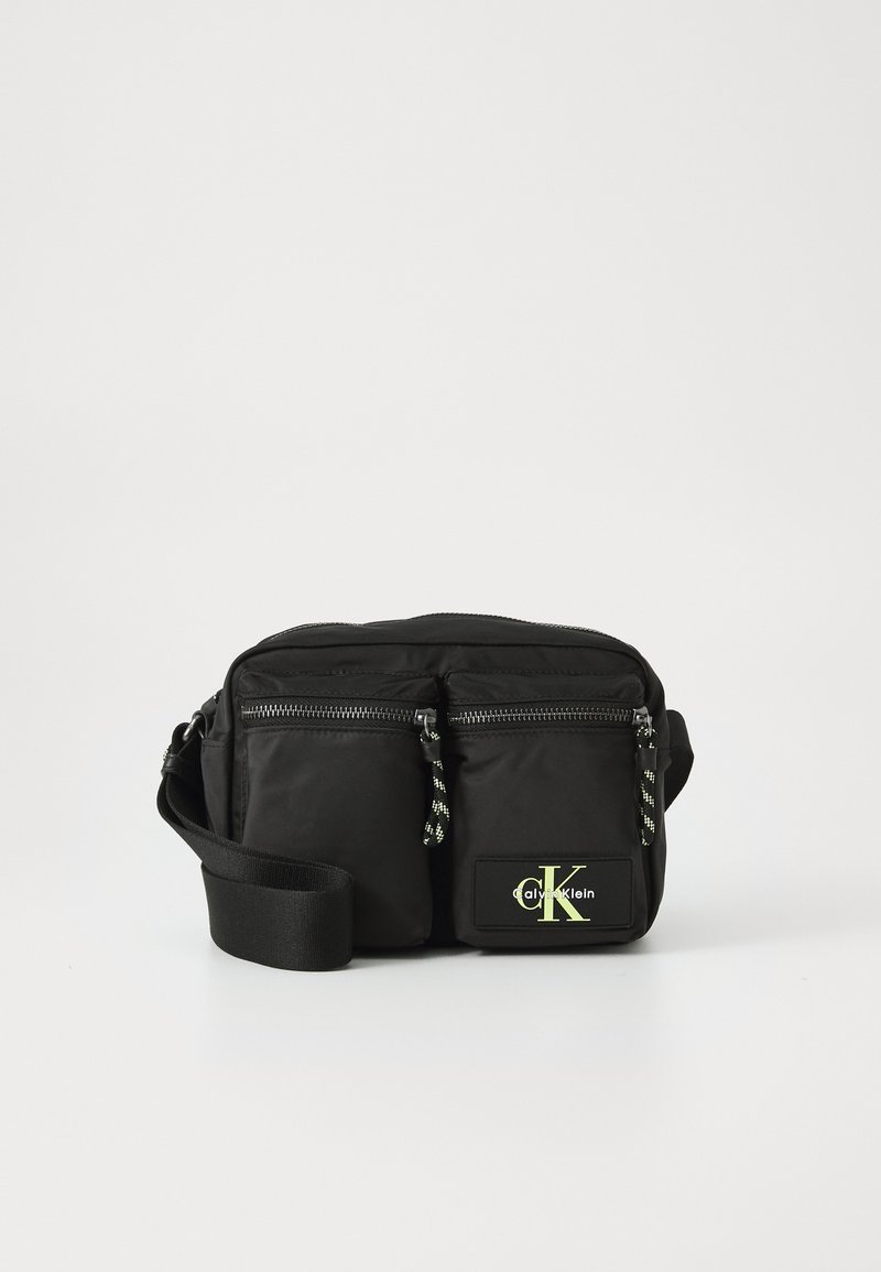 Black nylon crossbody bag with two front zip pockets, adjustable strap, and a yellow logo patch. Features a sleek and minimalist design.