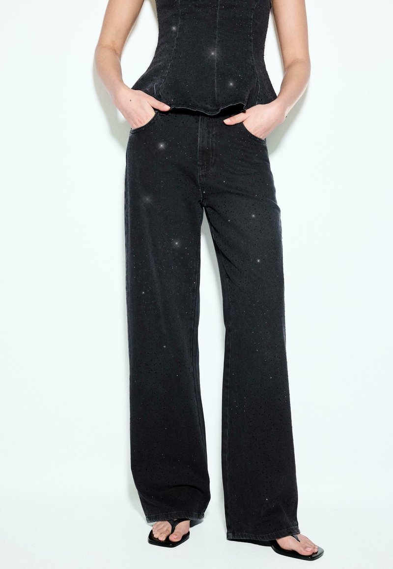 Femme portant un pantalon noir à jambes larges et un corset noir assorti avec des détails subtils scintillants, mains dans les poches, sandales noires visibles.