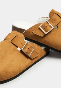Mules de ante marrón con un suave forro de piel blanca, hebillas de plata ajustables y una suela de goma negra texturizada. Diseño de punta redondeada.
