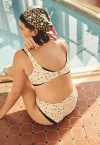 Floral bikini with a light cream background, black accent trim, and a clasp at the back. Model wears a patterned cap. Tile flooring.