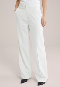 Pantaloni bianchi a gamba larga con chiusura a bottone, tasche laterali e una trama morbida e liscia. Abbinati a scarpe nere a punta.