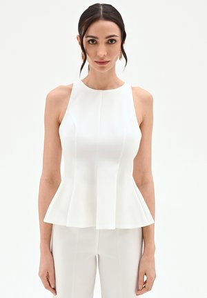 Femme aux cheveux foncés et boucles d'oreilles en or porte un haut péplum blanc sans manches et un pantalon blanc assorti sur un fond uni.