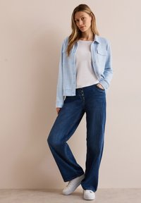 Lichtblauwe textuur knoopjack met twee voorzakken, gecombineerd met donkerblauwe wijde jeans en witte sneakers.