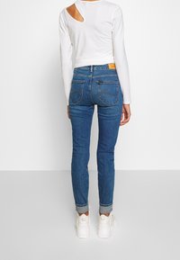 Jeans de denim azul de cintura alta con un ajuste Slim, que cuentan con bolsillos traseros y un desvanecimiento sutil, combinados con una blusa blanca con corte en los hombros.