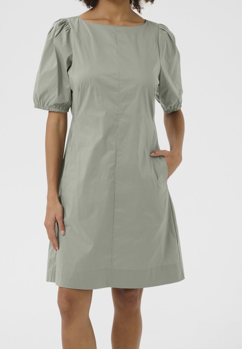 Ljusgrön, knä-length klänning med fyrkantig halsringning, puffade korta ärmar och sidofickor; gjord av mjukt, lätt tyg.
