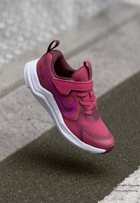 Nike športni čevelj v živo rožnati barvi z vijoličnimi poudarki, mrežastim zgornjim delom, zavezovanjem z vezalkami in oblazinjeno belo podplato, lebdeč nad sivo površino.