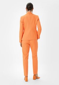Oranges Anzug mit einem taillierten Blazer und maßgeschneiderten Hosen, gefertigt aus einem glatten Stoff. Der Blazer verfügt über eine Rückennaht, und die Hosen sind schmal geschnitten.