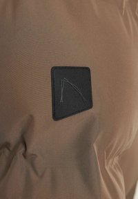 Tela marrón con una textura suave, con un parche rectangular negro que muestra un diseño minimalista. Detalles en relieve en el parche.