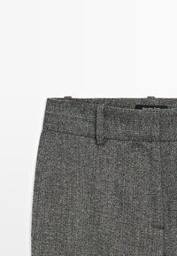 Szary teksturowany materiał paska spodni dopasowanych z szlufkami na pasek oraz częściowym widokiem metki na białym tle.