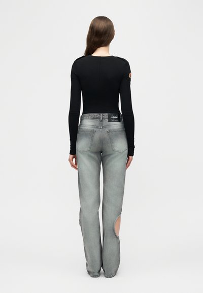 Γυναίκα με μακριά μαλλιά, που φοράει μαύρη μπλούζα μακρυμάνικη και γκρι τζιν ψηλόμεσο με κυκλικές περικοπές στις γάμπες πίσω, που στέκεται γυρισμένη με την πλάτη.