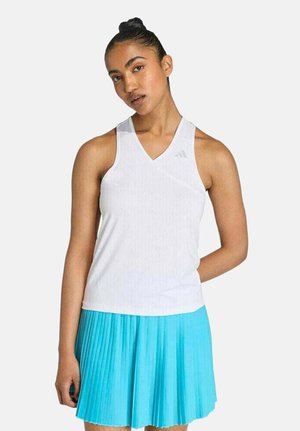 Mujer con una blusa blanca sin mangas de escote en V y una falda azul plisada, de pie con una mano detrás de la espalda contra un fondo liso.