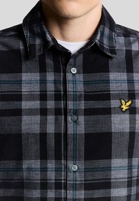 Camisa de cuadros negros y grises con cuello abotonado, que presenta un logo bordado amarillo y una tela texturizada con botones en contraste.