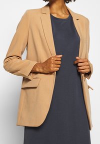 Blazer beige chiaro con vestibilità su misura, rever a punte, tasche laterali, tessuto testurizzato, indossato sopra un abito blu scuro. Nessun bottone visibile.
