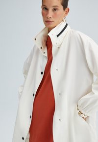 Giacca impermeabile bianca con colletto alto, dettagli con zip nera e bottoni a pressione. Indossata sopra un top color ruggine con polsini elastici.