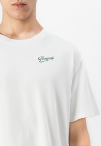Close-up of a person wearing a white T-shirt with green "Pompeii" text logo on the upper left chest against a plain white background.